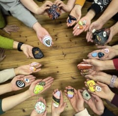 Useat kädet muodostaa ympäyrän pöydän päälle. Jokaisessa kädessä on maalattu kivi.