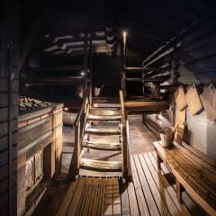 Sisäkuva savusaunasta, iso kiuas vasemmalla, alapenkit lattiatasossa ja portaat varsinaisille lauteille.