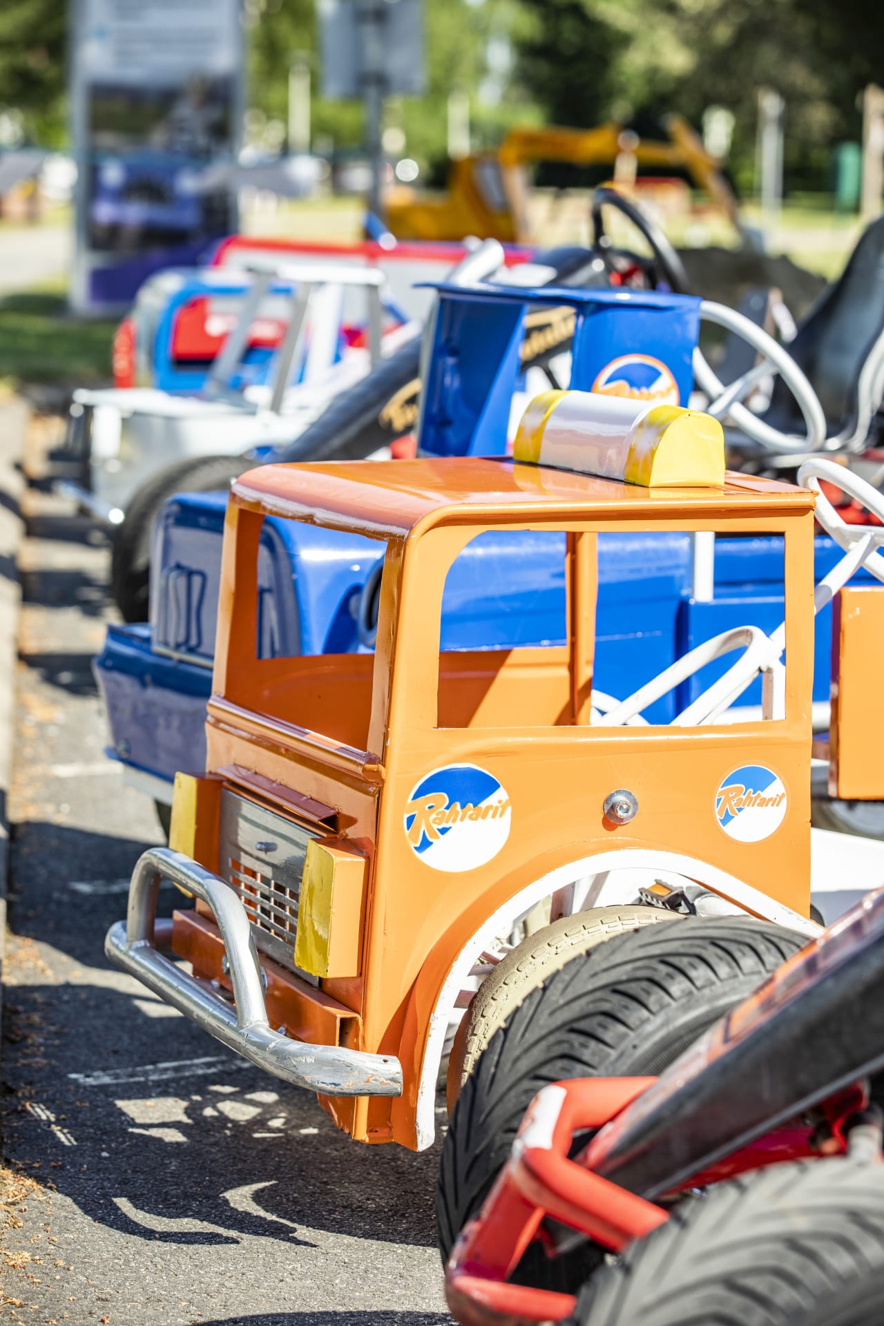 Liikennepuiston polkuautoja parkkirivistössä. Autot ovat erimallisia ja erivärisiä, kuvan etualalla oranssi ja sininen polkuauton keula.
