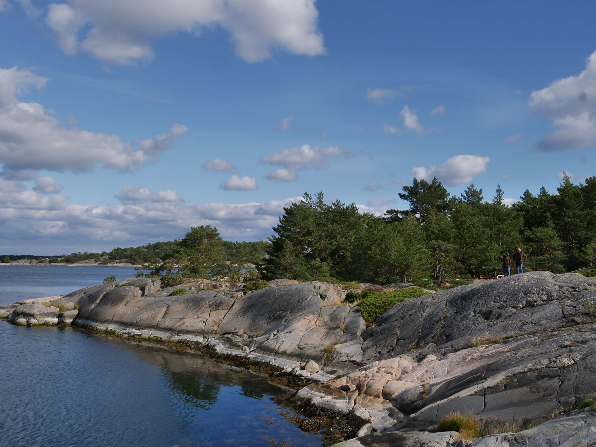 Kuvassa on jyrkkää kalliorantaa. Kalliolla kävelee kaksi ihmistä. Ihmisten takana kasvaa puita.The picture shows a steep rocky shore. Two people are walking on the rock. Trees grow behind people.