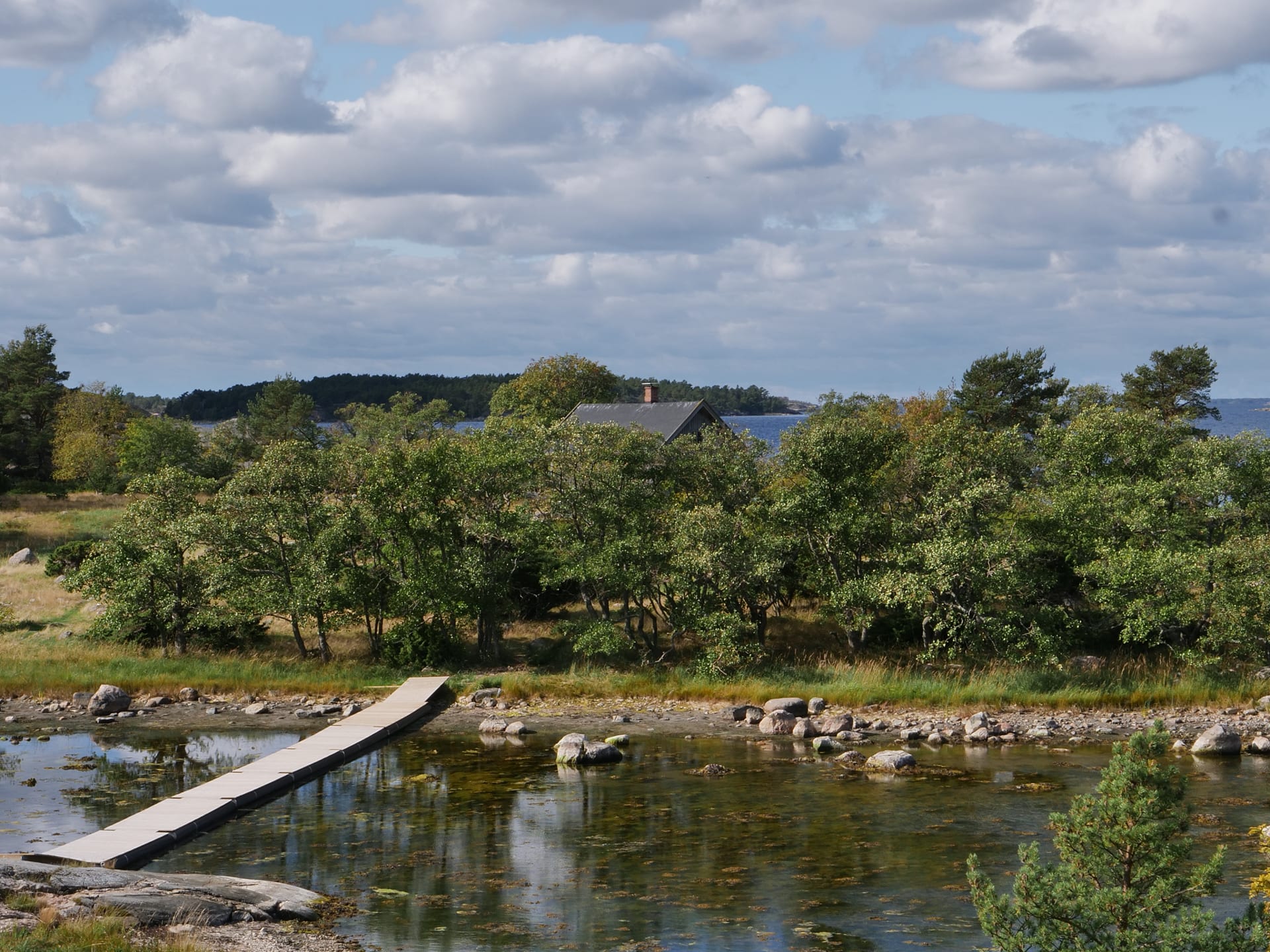  Vasemmalla reunalla näkyy seuraavalle saarelle vievä ponttonisilta. Saaren puuston yli pilkottaa talon katto.The picture is taken from a high rock down. On the left side you can see the pontoon bridge leading to the next island