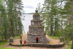 Neljä retkeilijää kivistä rakennetulla muistomerkillä. Taustalla on järvi. For hikers are beside a monument which is built of stones. In the background there is a lake.