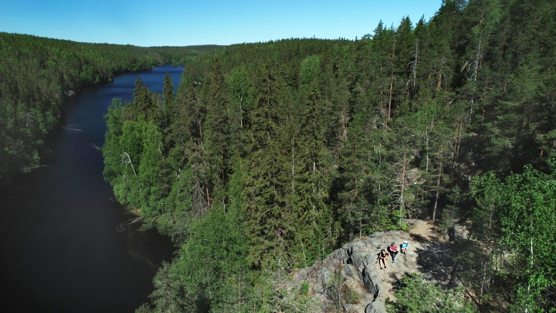 Ilmakuva kapeasta järvestä, jonka oikealla puolella kohoaa korkea kallio, muutoin järvi on metsien ympäröimä. Kalliolla istuu kolme retkeilijää. Kuva: Jari Salonen.
