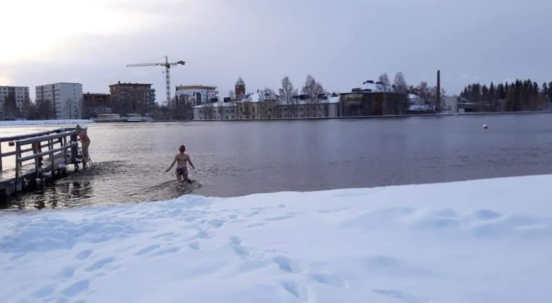 A person wading in ice cold water in Tuira beach in winter. Foreground snow in the bank. Background buildings on the other side of the Oulu river. Sky is grey and clouded.