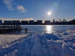 A person dipping in the Oulu river in winter in Tuira beach. Foreground snow pile. Background buildings on the other side of the river. Sun is shining.