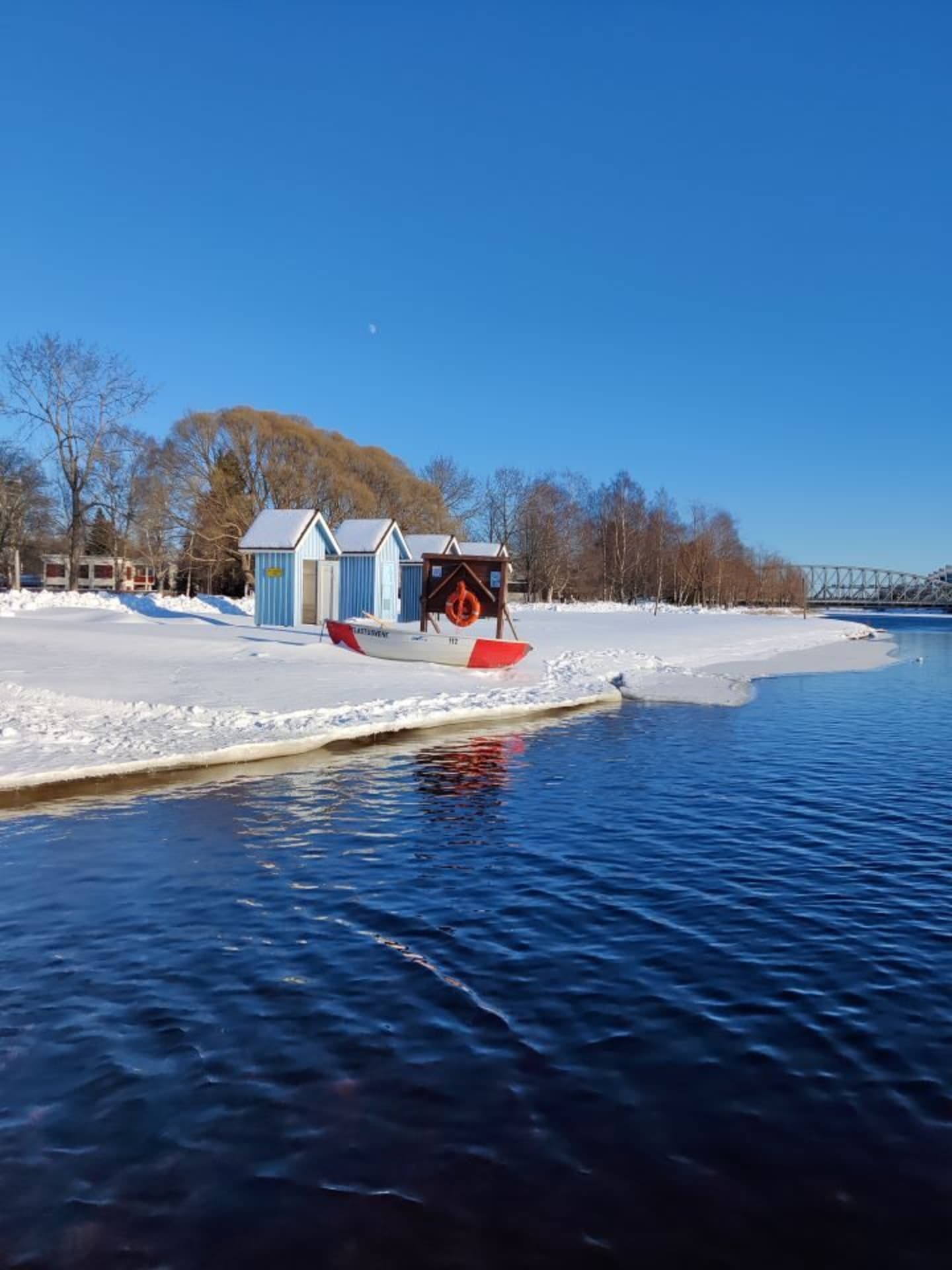 Light blue bathing huts in Tuira beach. Foreground blue Oulu river. Snow on the bank. Background brown trees without leaves.