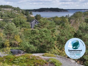 Kuva on otettu kallion laelta yli saaren. Harmaa talo erottuu mäntyjen keskellä. Kuvan takaosassa on merta ja muita saaria. The gray house stands out among the pine trees. In the back of the picture is the sea and other islands.