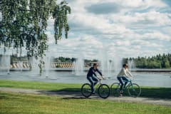 Two persons biking in Oulu river delta area in summer. Foreground green grass. Background fountains in Oulu river and dam of the Oulu hydro electric power plant.