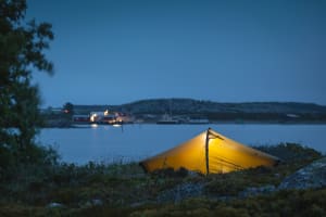 On yö. Etualalla on sisältä valaistu teltta. Teltan jälkeen on merta, jonka toisella puolelta erottuu sataman valoja.It is night. In the foreground is a tent lit from the inside.