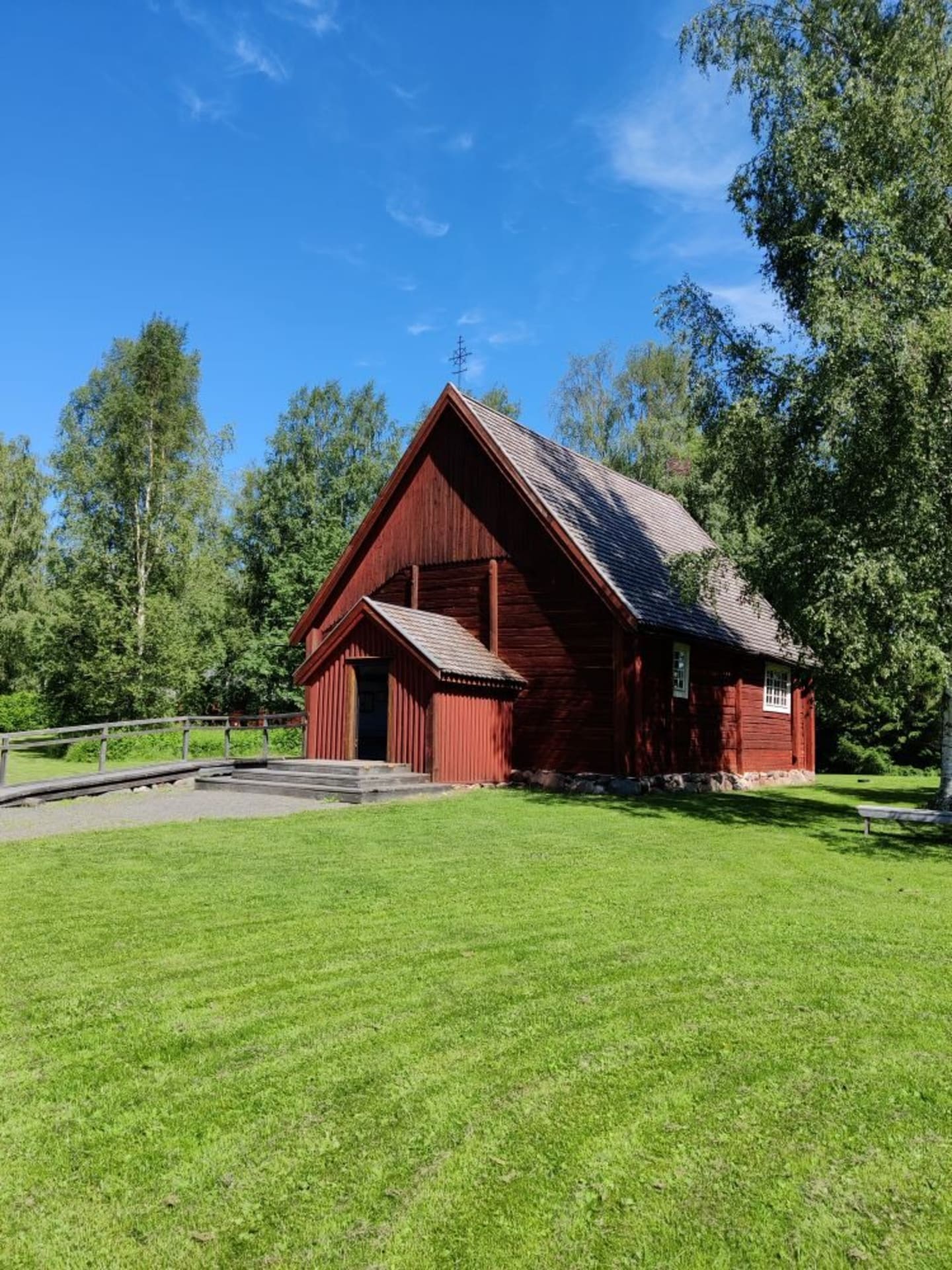 The old red wooden church in Turkansaari in summer. Foreground green grass, background green birch trees.