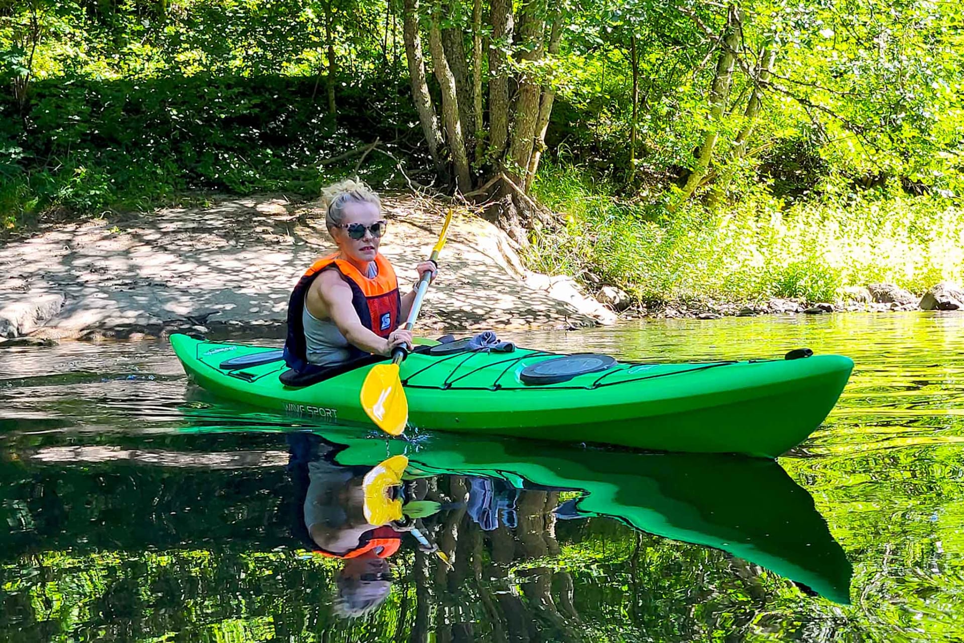 A lady in a green Kayak paddling on river. Photo is taken on a front. You can see her face.