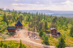 Taukopaikan pihapiiriä kesäisessä Pallas-Yllästunturin kansallispuistossa. Etualalla puuvaja, kota, kuivakäymälä ja nuotiopaikka. Taustalla metsää ja tuntureita. Kuva: Rami Valonen