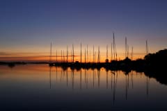 Aurinko värjää horisontin oranssiksi. Horisontin edessä kuvan keskellä on veneitä mustina. Tyyni vesi tumman sinistä. Kuva: Jarmo Vehkakoski.