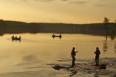 Kaksi kalastajaa. Taustalla tyyni järvi ilta-auringossa ja järvellä kaksi soutuvenettä. Kuva: Ismo Pekkarinen