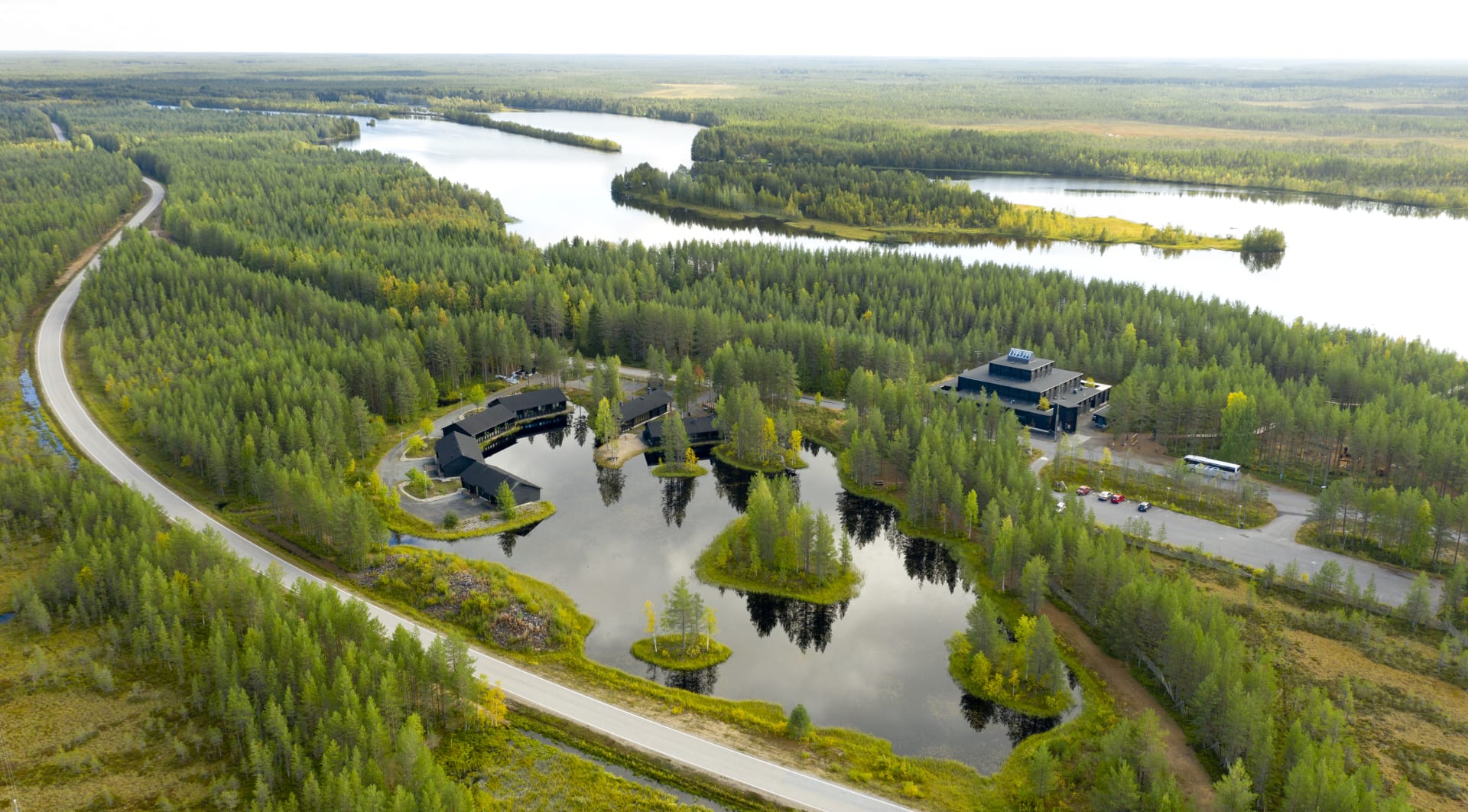 Ilmakuva, iso hirsirakennus lammen rannalla, metsäisessä maisemassa, taustalla joki.