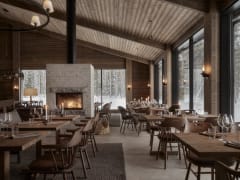 A cozy restaurant dining room at Jänkä Resort with wooden tables, soft lighting, candles, and a fireplace, set against floor-to-ceiling windows overlooking a snow-covered forest.