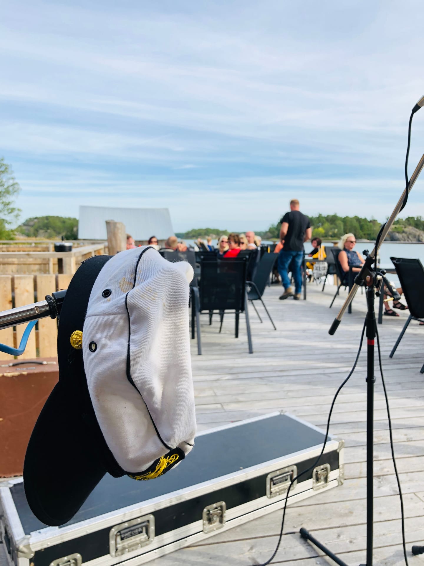 A captain's hat perched on a microphone stand sets a maritime theme at an outdoor performance, with the audience in the background enjoying the ambiance at Cafe Laituri.