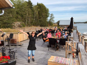 A cheerful person with arms wide open stands at the forefront of a lively outdoor venue by the water, with guests seated at tables enjoying the ambiance.