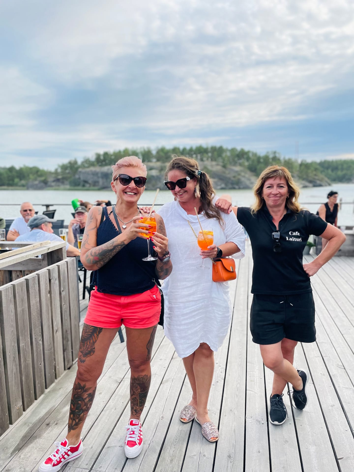 Two guests, enjoying their drinks, share a cheerful moment with a staff member on a lively deck, capturing the joy of socializing by the waterside.