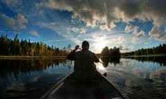 Yksinäinen meloja metsien ympäröimällä lammella, pilvet heijastuvat lampeen.