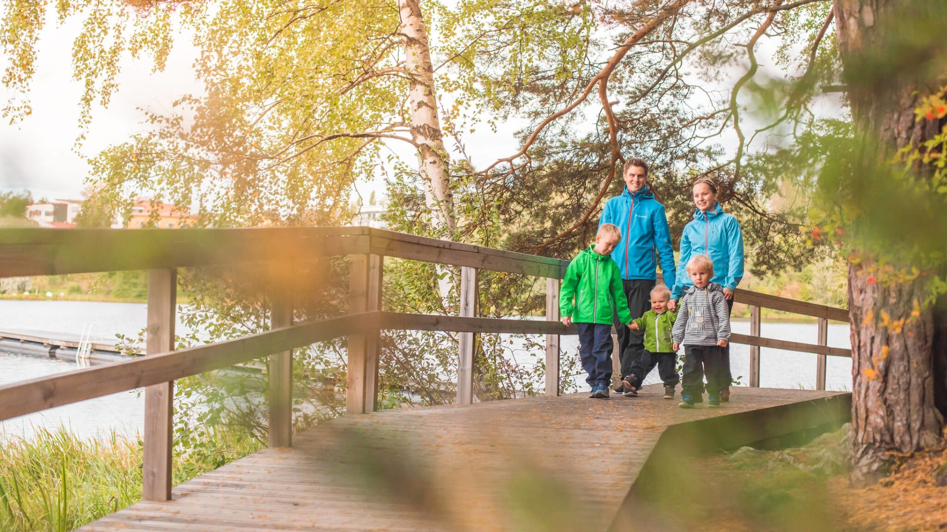 Perhe kävelee puista siltaa pitkin järven rannassa.