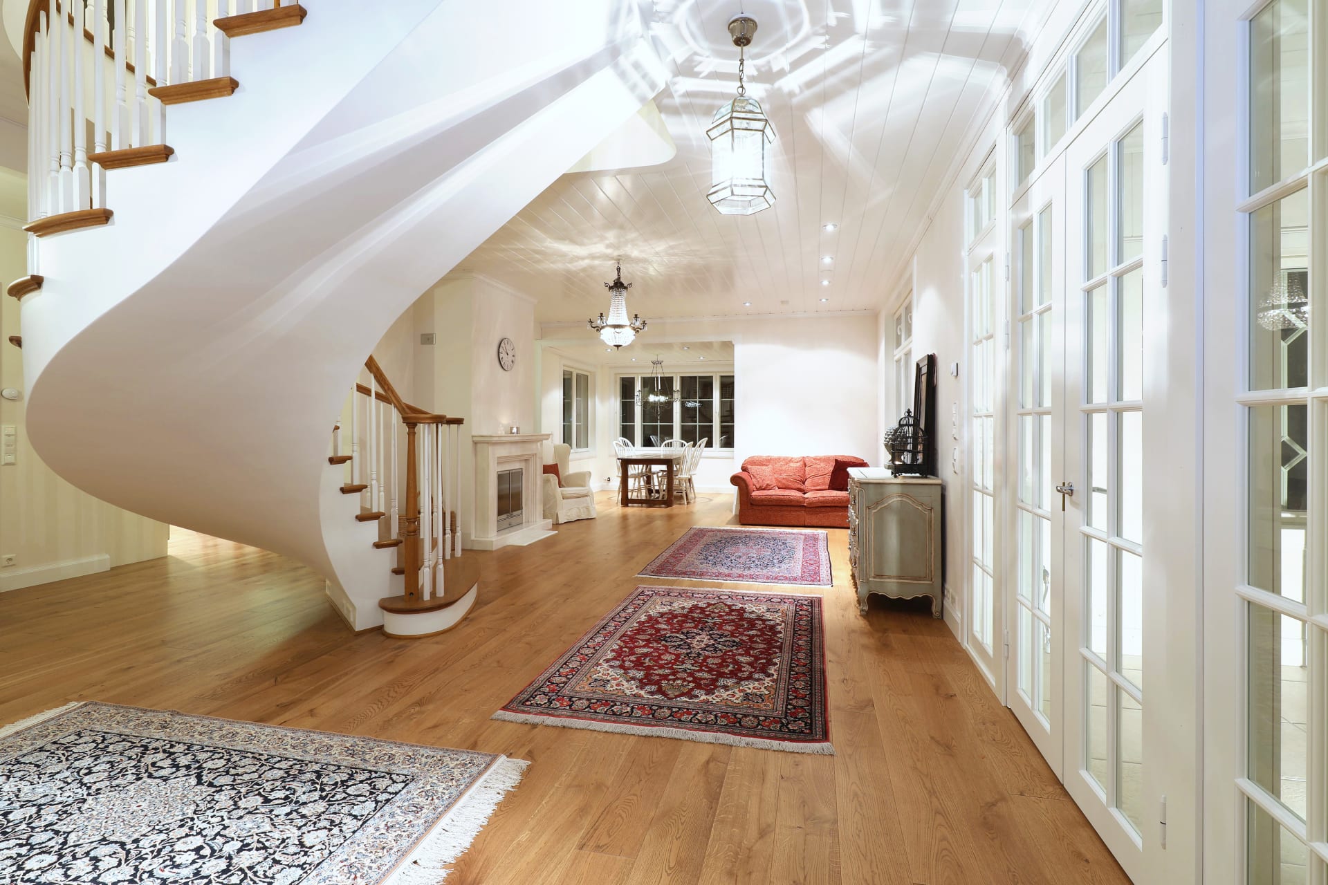 A view over the oak parquet of Villa Cone Beach living room, showing curved stairs and a white marble fireplace, seen from the TV room to the dining area.