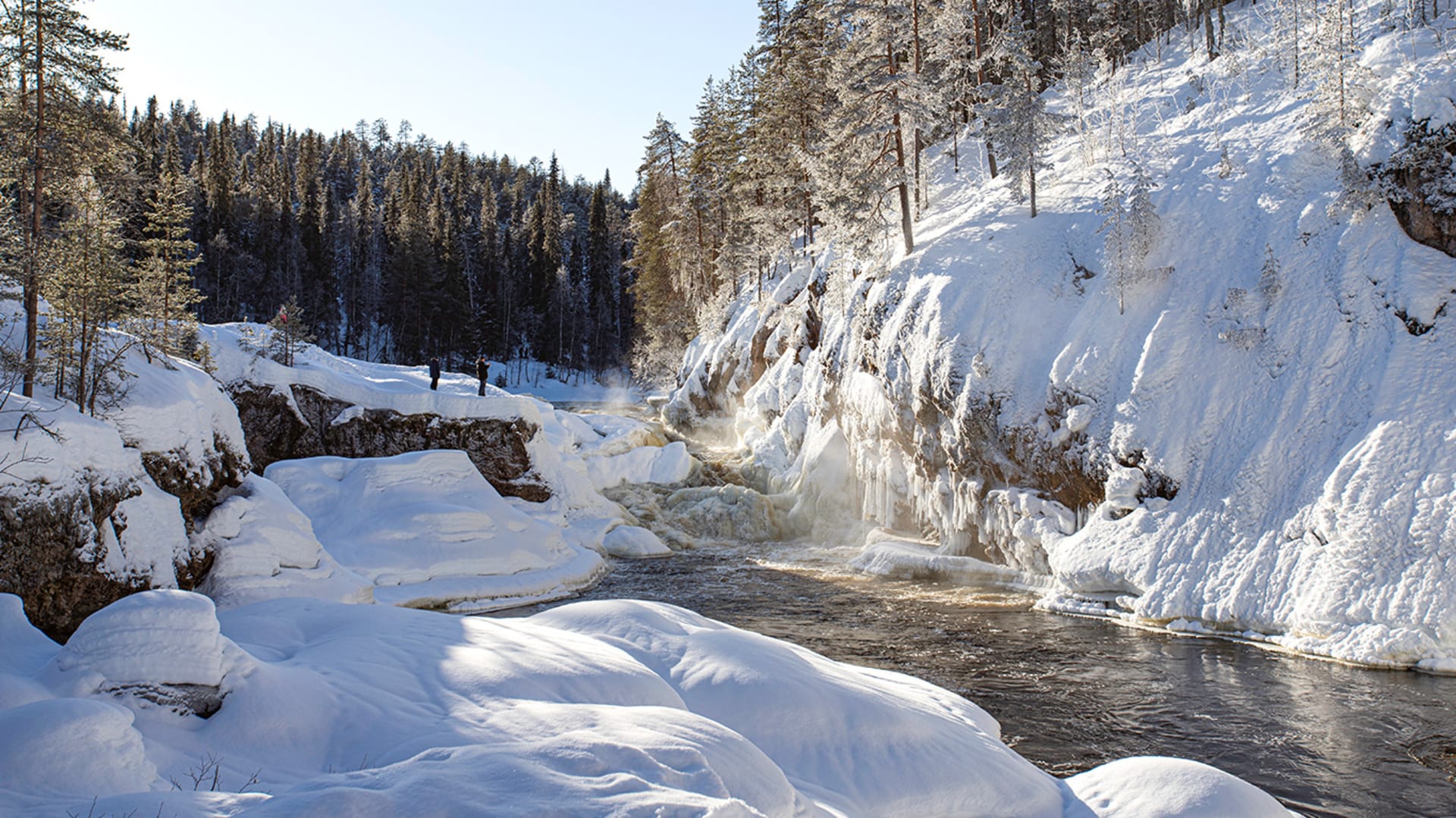 Luminen koskimaisema. Retkeilijät seisovat ihailemassa kuohuavaa koskea. /  Snowy rapids landscape. Hikers stand admiring the bubbling rapids. Kuva: Minna Koramo