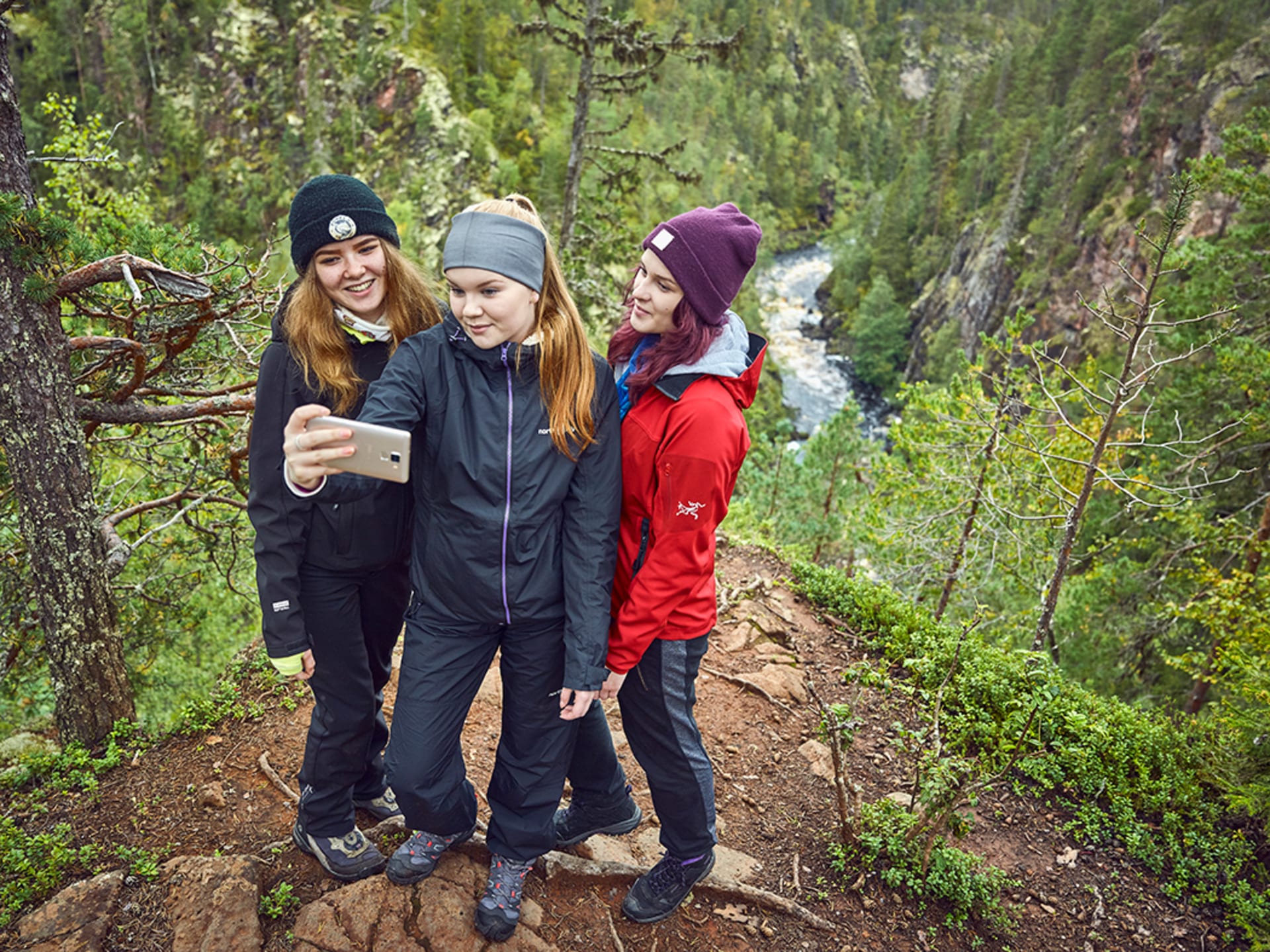 Kolme retkeilijää ottaa kuvaa kallioseinämän päällä. Takana avautuu rotkolaakso, jonka pohjalla virtaa joki. / Three hikers take a picture on top of a rock wall. Behind opens a gorge valley, at the bottom of which flows a river. Kuva: Jari Hindström