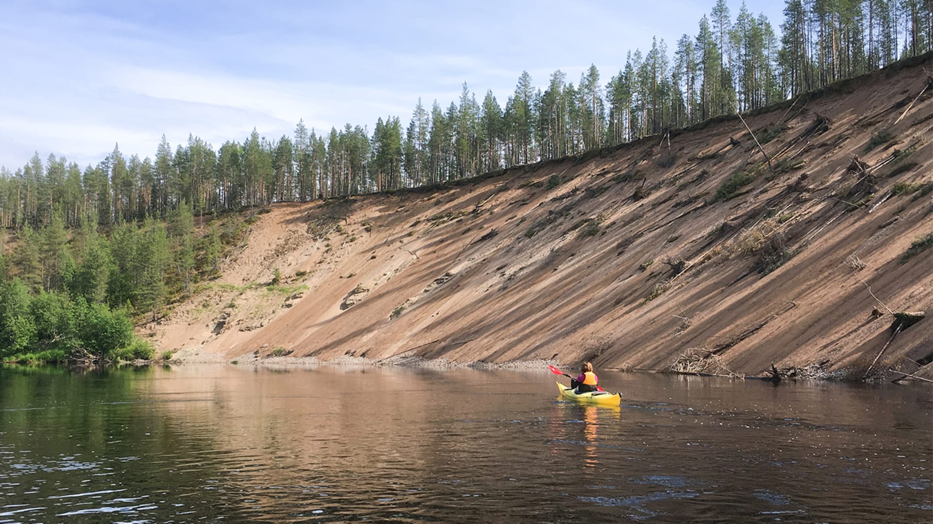 Meloja valtavan suoraan joesta kohoavan hiekkarinteen vierellä. Ympärillä kesäinen maisema. /  Paddle next to a huge sandbank rising directly from the river. Summer landscape all around. Kuva: Minna Koramo