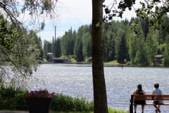 Järvimaisema. Rannalla penkki, jossa istuu kaksi ihmistä maisemaa katsellen.