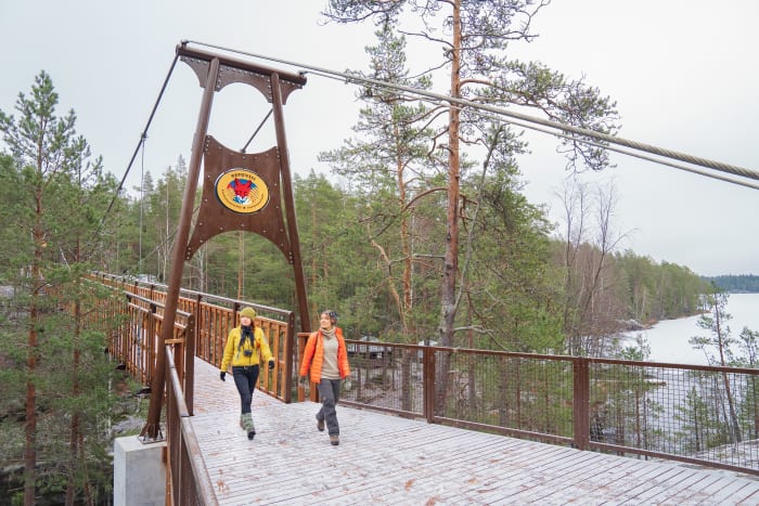 Kaksi retkeilijää kävelee puukantisella sillalla. Maassa on ohut kerros lunta ja taustalla näkyy jäätynyt järvi. Kuva: Jouni Kallio