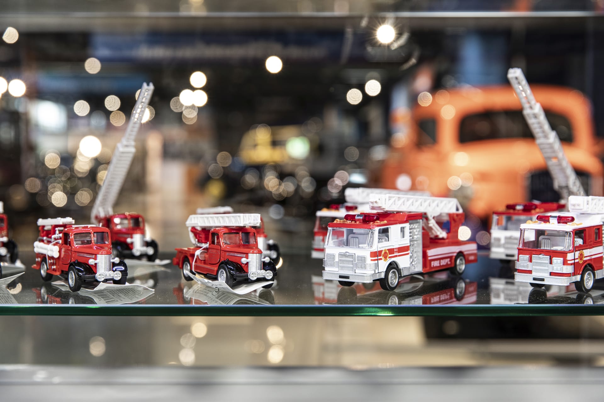 Pieniä leikkipaloautoja lasihyllyllä, takana näkyy epäselvinä hahmoina isoja näyttelyautoja.