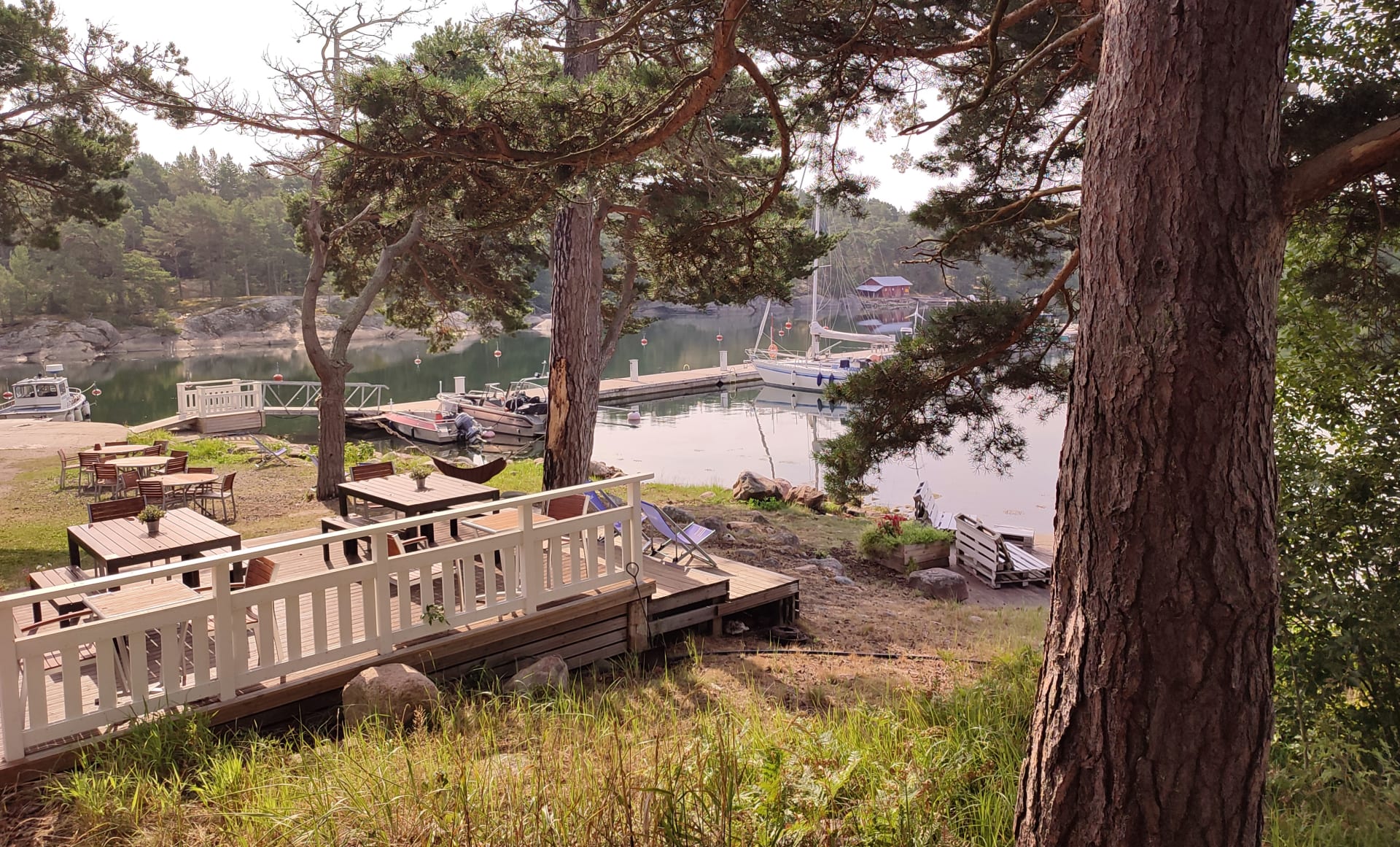 Kuva on otettu saarelta kohti laituria. Väliin jää terassi, jossa on pari pöytää sekä puiden väliin kiinnitetty riippukeinu. picture was taken from the island towards the pierce couple of tables and a hanging swing attached between the trees.