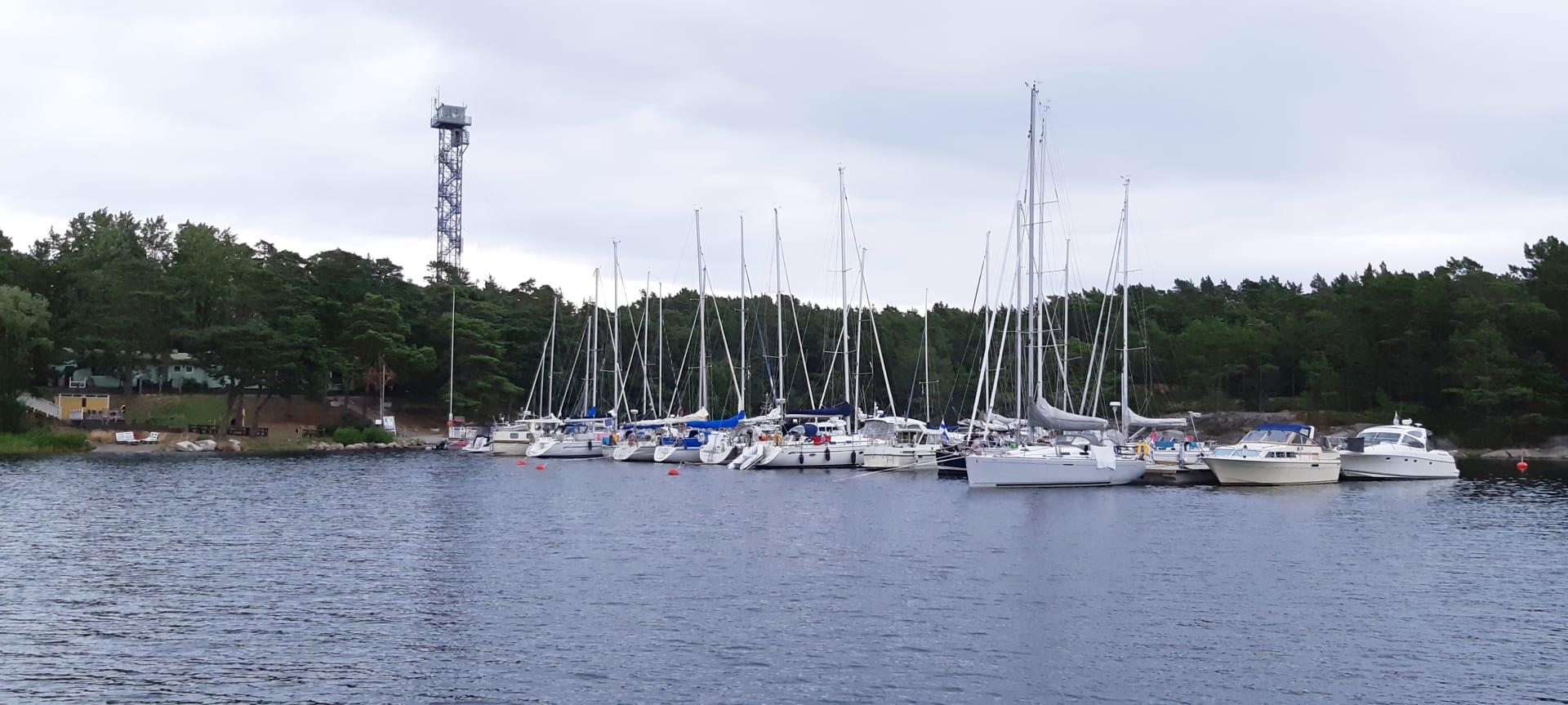 Pitkällä laiturilla on paljon veneitä. Rannalla  rakennus sekä korkea torni. The picture was taken from the water. There are many boats on the long pier. A building and a tall tower stand out on the beach. There are many trees on the beach.