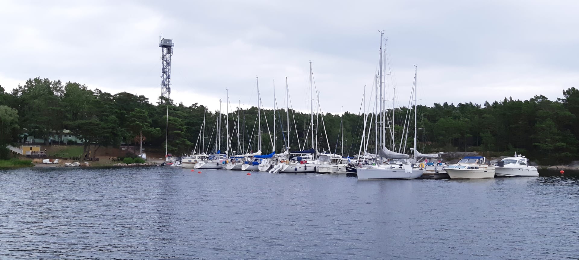 Pitkällä laiturilla on paljon veneitä. Rannalla  rakennus sekä korkea torni. The picture was taken from the water. There are many boats on the long pier. A building and a tall tower stand out on the beach. There are many trees on the beach. Pitkällä laiturilla on paljon veneitä. Rannalla  rakennus sekä korkea torni. The picture was taken from the water. There are many boats on the long pier. A building and a tall tower stand out on the beach. There are many trees on the beach.
