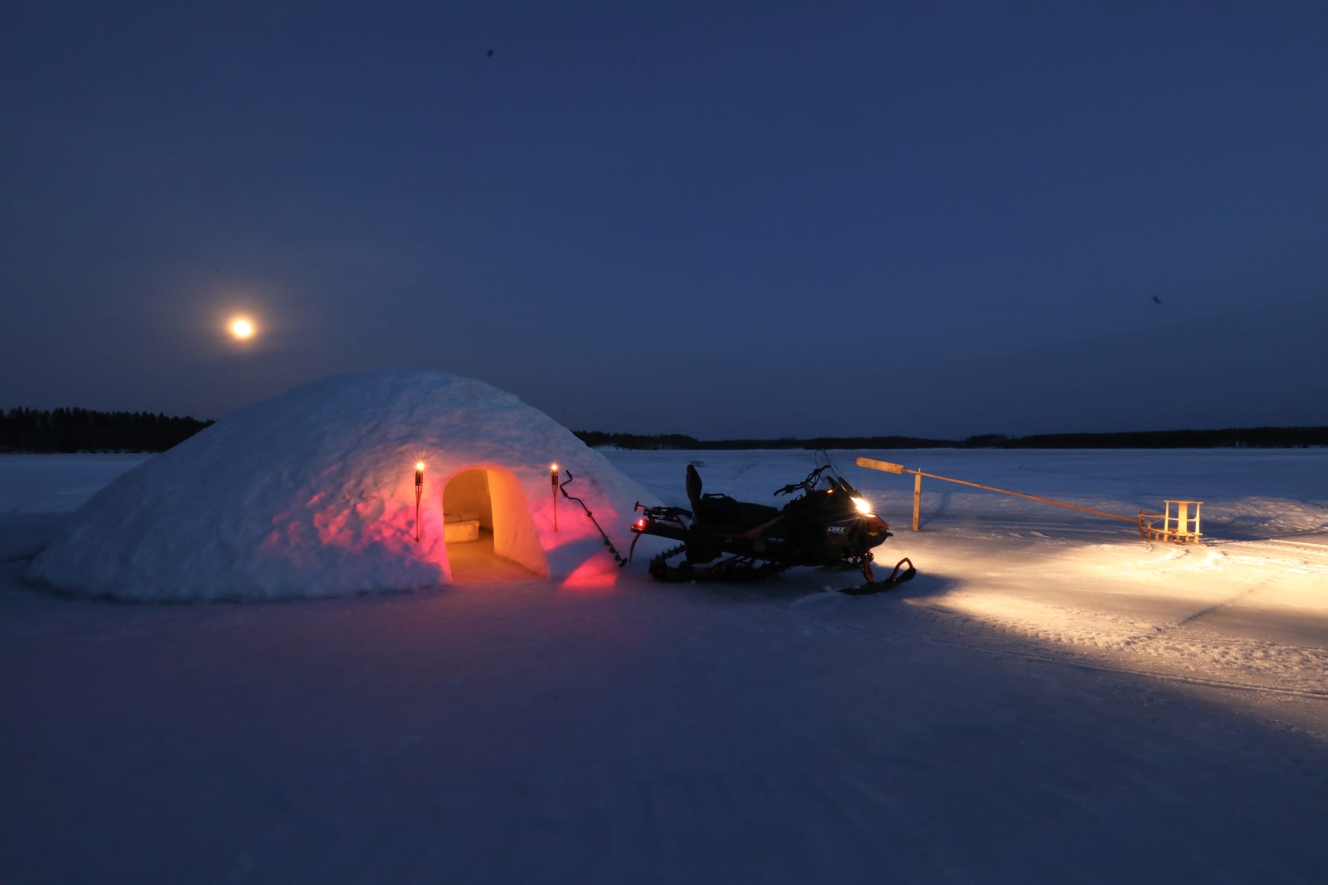 A snow igloo, merry-go-round sled, and snowmobile set on lake ice under a full moon in the blue dusk.