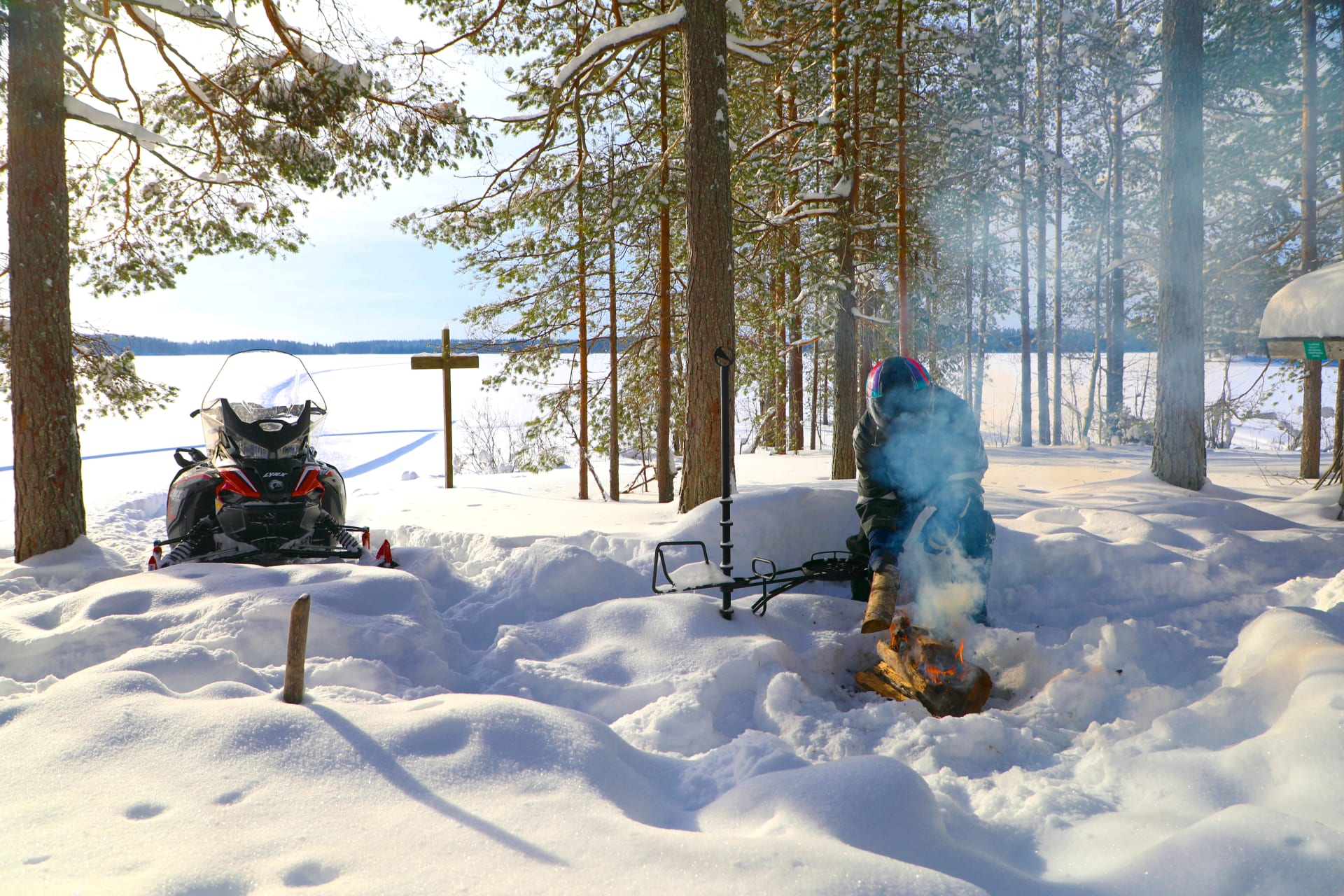 A snowmobile driver has left tracks on virgin snow across a lake and is lighting a campfire to rest.