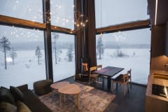 A cozy villa living and dining area at Jänkä with a fireplace, wooden furniture, soft lighting, and floor-to-ceiling windows overlooking a snowy landscape.