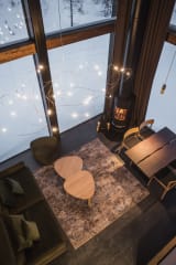 An open-plan villa lounge and dining area at Jänkä with wooden furniture, soft lighting, a fireplace, and large windows facing the winter forest.