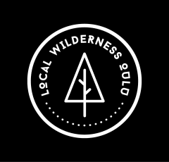 Local Wilderness Oulu logo. White logo on black background. A circle that has a tree in it and the text Local Wilderness Oulu.