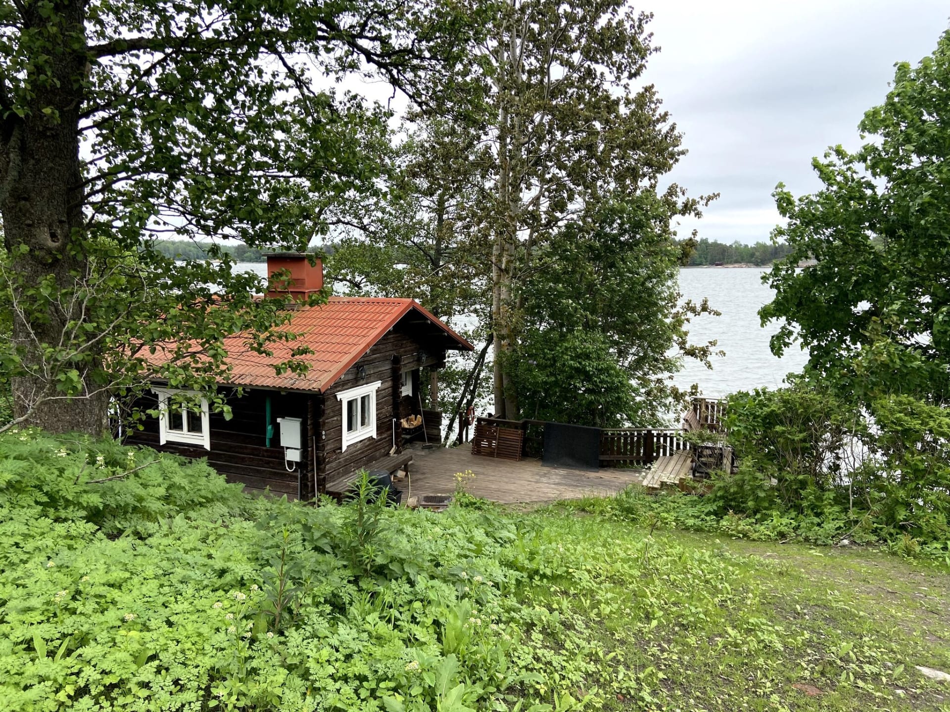 Wooden sauna building by the sea, surrounded by trees, with a terrace and stairs leading to the shore.
