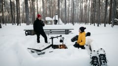 Kaksi lumikenkäilijää tauolla lumisessa metsässä. Toinen on polvistunut nuotion ääreen. Taustalla kota ja halkovaja. Kuva: Eeva Mäkinen