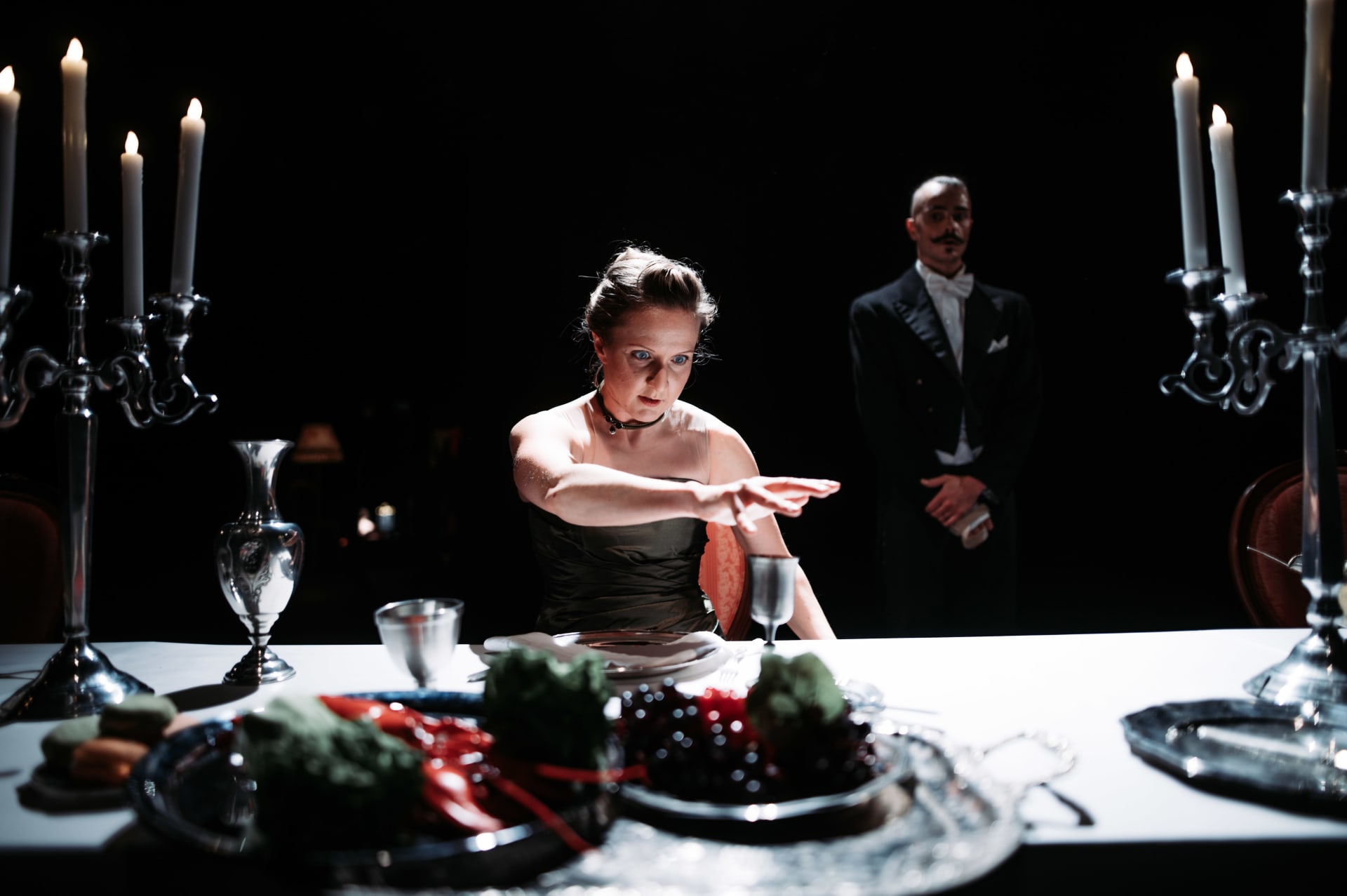 A woman in a black dress sits at a dinner table, with a man in a tuxedo standing behind her. The table is set with candles, glassware and food. From Aurinkobaletti’s dance theatre production Illallisvieraita.