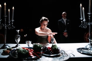 A woman in a black dress sits at a dinner table, with a man in a tuxedo standing behind her. The table is set with candles, glassware and food. From Aurinkobaletti’s dance theatre production Illallisvieraita.