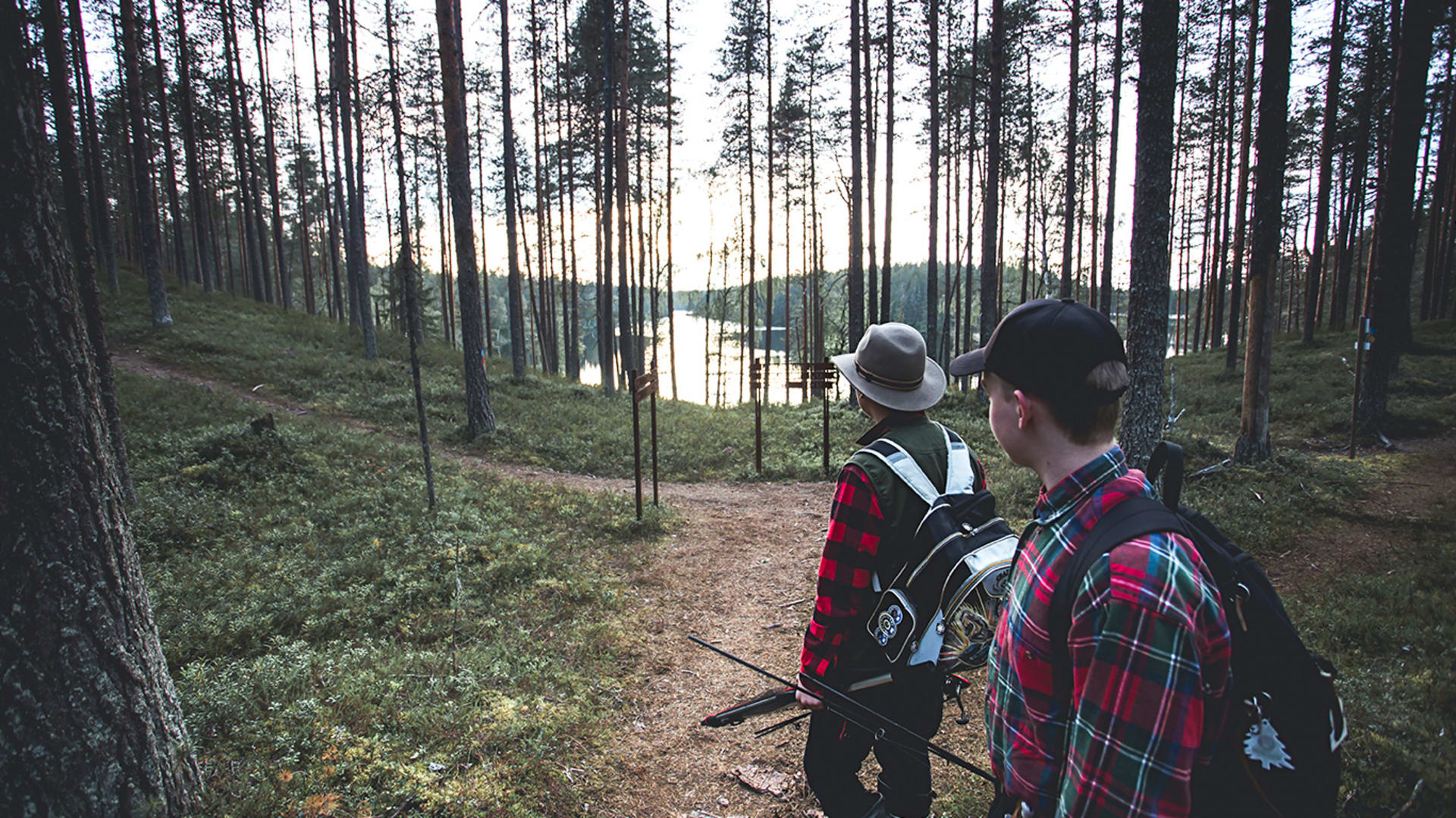 Kaksi kalastajaa polulla kesäisessä luonnssa. Ympärillä mäntymetsää ja metsän läpi siintää järvi. / Two fishermen on a path in summer nature. There is a pine forest around and a lake through the forest. Kuva: Maarit Vaahteranoksa