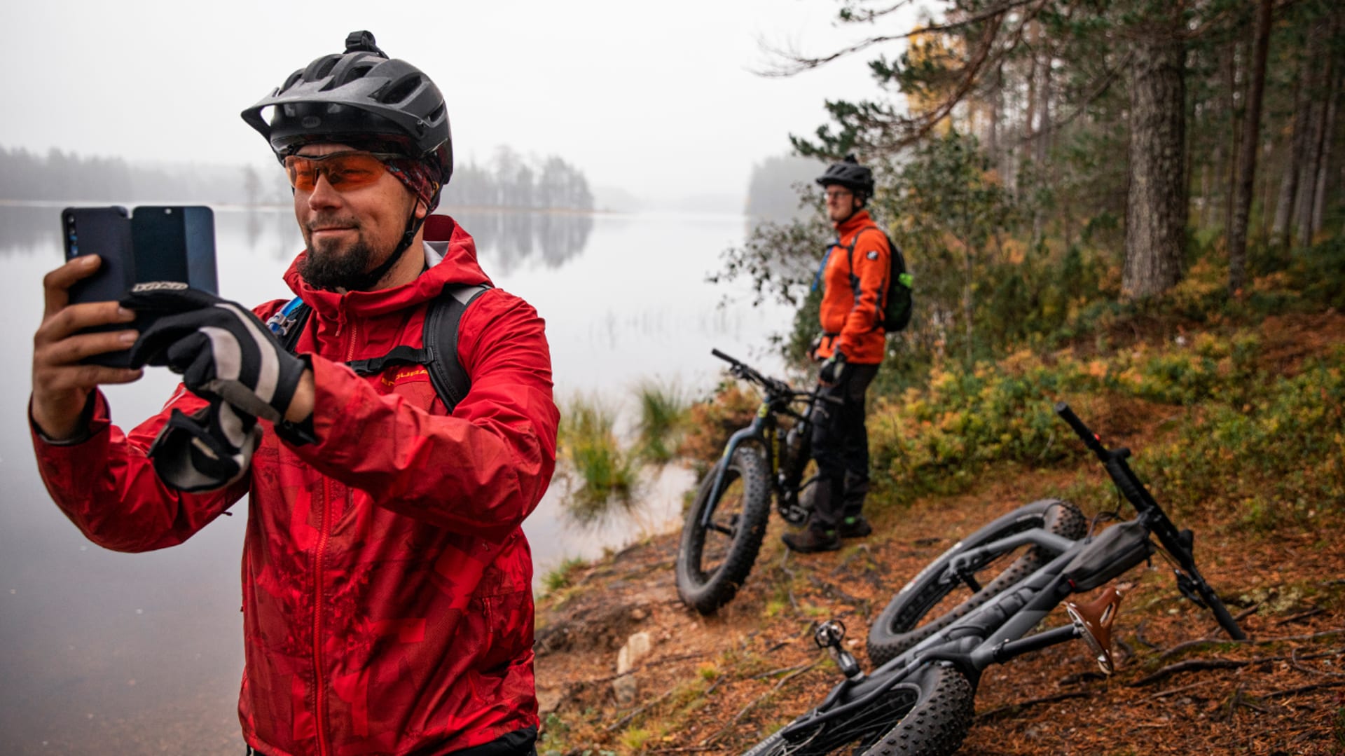 Etualalla kypäräpäinen mies ottamassa valokuvaa kännykällä järven rannalla. Taustalla toinen pyöräilijä. / In the foreground, a man with a helmet taking a photo with a mobile phone by the lake. Another cyclist in the back. Kuva: Harri Tarvainen