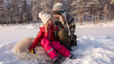 Lapsi ja aikuinen lumisellä järvenjäällä pilkillä. Lapsi istuu lampaantaljan päällä. / A child and an adult with ice on a snowy lake ice. The child is sitting on a sheepfold. Kuva: Jonna Kalliomäki.