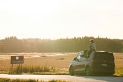 A couple on a roadtrip on the Taikayöntie Road. They are sitting on the roof of a van, admiring a field in the sunshine.
