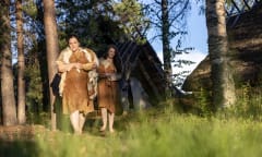 two people in leather clothes, barefoot, in front of a Stone Age dwelling