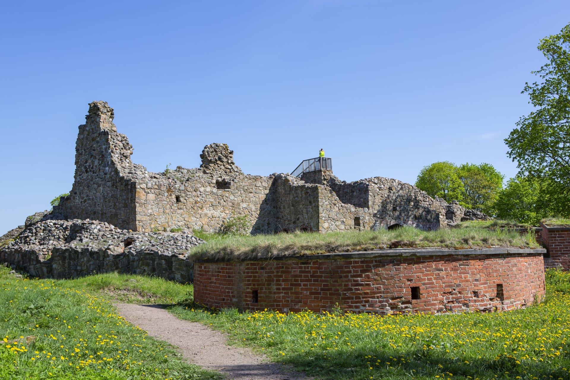 Taustalla näkyy linnanraunioiden matalia kiviseiniä. Raunioiden päällä näkyy ihmisiä näköalatasanteella. Etualalla pyöreä punatiilinen matala rakennelma. On kesä, aurinko paistaa. Kuva: Jari Kostet Taustalla näkyy linnanraunioiden matalia kiviseiniä. Raunioiden päällä näkyy ihmisiä näköalatasanteella. Etualalla pyöreä punatiilinen matala rakennelma. On kesä, aurinko paistaa. Kuva: Jari Kostet